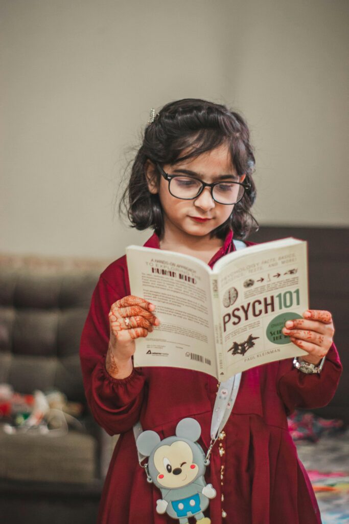 A young girl with eyeglasses and henna tattoos reading a book indoors.